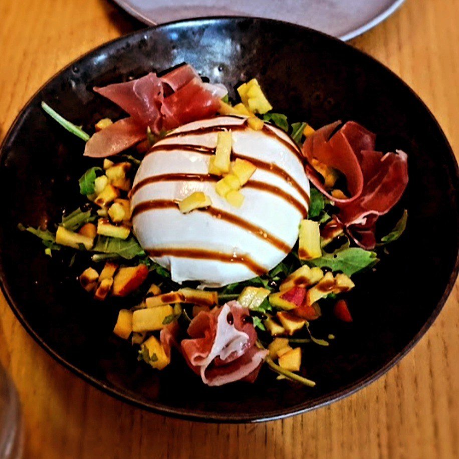 Burrata inteira com fio de redução escura, sobre folhas verdes, pêssego em cubos e fatias de presunto, numa taça preta